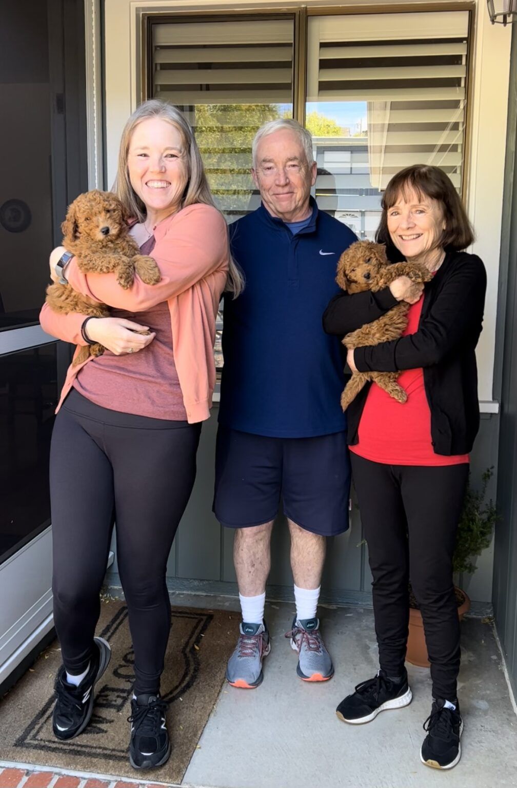 Three adults standing together outside with two small brown puppies.